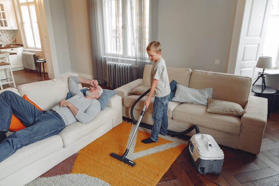 A person kneeling on a patterned carpet in a room, emptying a dustbin into a yellow portable vacuum cleaner with a black lid and a flexible hose. The carpet has a floral design with neutral tones. The