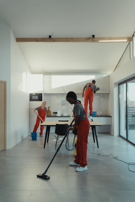 A person wearing orange rubber gloves is arranging a lightbox sign with the words 'CLEANING HOME' in bold, black letters. The lightbox is positioned on a wooden surface in a well-lit room, likely a ki