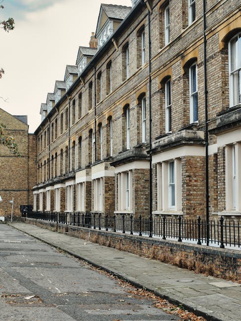A row of multi-storey Victorian-style residential buildings constructed with grey brick facades, each with large sash windows and peaked, slate-tiled roofs. The buildings are situated behind a grassy 