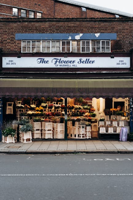 The exterior of The Flower Seller shop on Muswell Hill features a large, horizontal storefront with a white sign displaying the business name in elegant blue script. The upper part of the building is